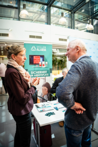 A woman and a man engage in conversation at a promotional booth for "Stiller Alarm" (Silent Alarm) during a trade fair. The woman gestures while speaking next to a banner that reads "SICHERHEIT AM ARBEITSPLATZ" (Safety at the Workplace), which shows software for computers and mobile devices. The setting is a bright, modern atrium with large glass windows.